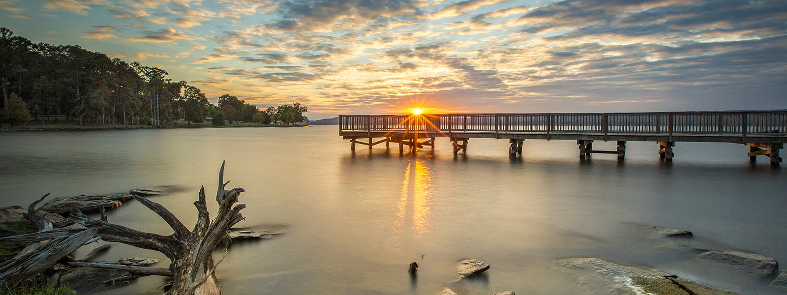 Sunset over Lake Guntersville, Alabama — a wooden fishing pier stretches across still water as the sun sets through a dramatic sky. Photo by John Sharp.